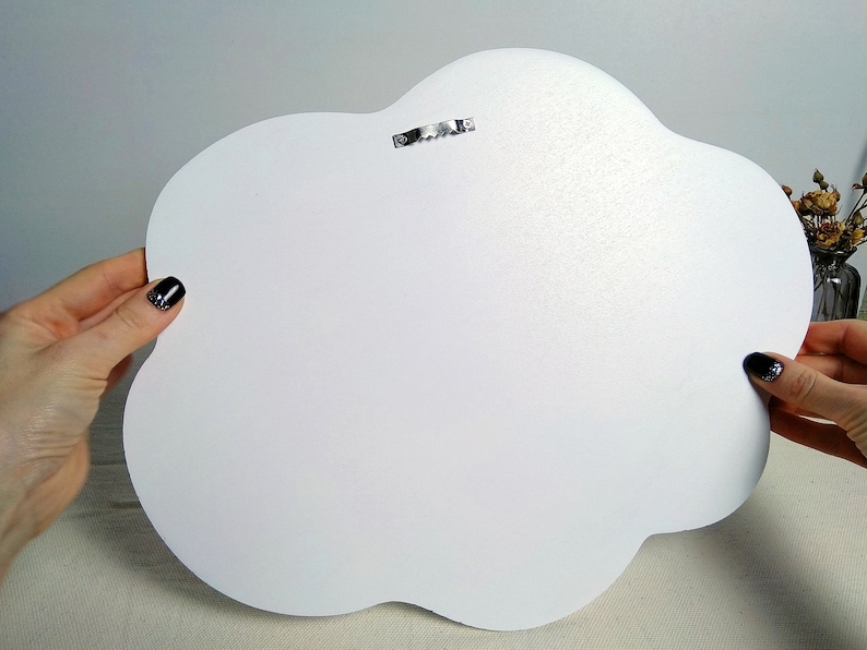May include: A white cloud-shaped wooden wall decoration with a metal sawtooth hanger on the back. The cloud has a smooth, matte finish. The image shows the back of the cloud, held by hands with dark nail polish.