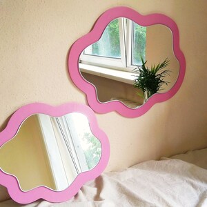 May include: Two cloud-shaped mirrors with pink frames. The mirrors are mounted on a light beige wall. The mirrors reflect a window and a plant. One mirror is mounted on the wall, and the other is resting on a white sheet.