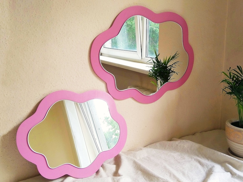 May include: Two cloud-shaped mirrors with pink frames. The mirrors are mounted on a light beige wall. The mirrors reflect a window and a plant. One mirror is mounted on the wall, and the other is resting on a white sheet.