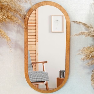 May include: A wooden oval-shaped mirror with a rounded top and bottom. The mirror is hanging on a white wall with a textured pattern. There is a chair and a picture frame reflected in the mirror.