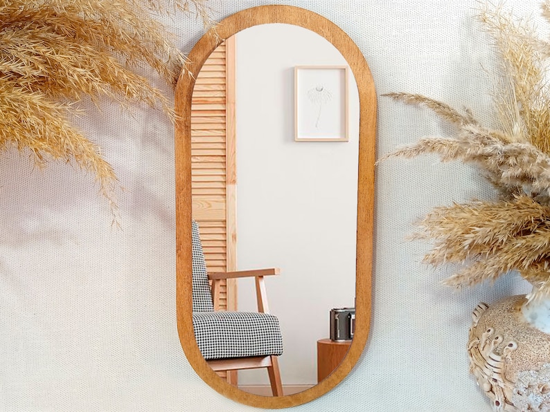 May include: A wooden oval-shaped mirror with a rounded top and bottom. The mirror is hanging on a white wall with a textured pattern. There is a chair and a picture frame reflected in the mirror.