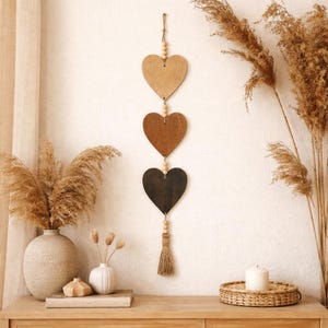 May include: A wooden wall hanging featuring three heart-shaped cutouts in varying shades of brown, suspended by a string with wooden beads. A tassel hangs from the bottom heart. The decor is displayed against a white wall, with dried plants and a candle.