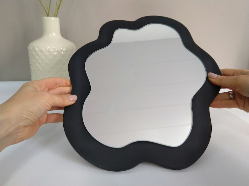 Pu&ograve; includere: Uno specchio nero di forma irregolare con una superficie liscia e riflettente. Lo specchio ha una cornice nera spessa.