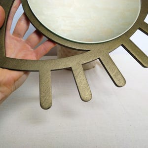 K&ouml;nnte beinhalten: Ein runder Wandspiegel mit einem metallisch bronzefarbenen Rahmen in Augenform, von dessen Rand strahlenf&ouml;rmige, wimpernartige Elemente ausgehen. Der zentrale Glasspiegel ist kreisf&ouml;rmig und reflektierend, w&auml;hrend der Rahmen matt ist.
