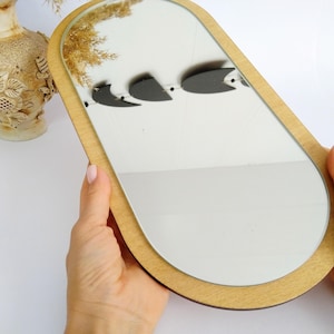 Op de afbeelding: Ovale spiegel met een licht houten frame. De spiegel reflecteert drie zwarte vogel silhouetten. De spiegel wordt tegen een witte achtergrond gehouden. Een decoratieve vaas is zichtbaar op de achtergrond. De spiegel is ongeveer 30 cm lang.