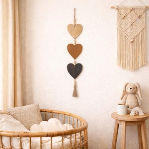 May include: A vertical wall hanging featuring three wooden heart-shaped cutouts in graduated shades of light, medium, and dark brown, connected by a string of small wooden beads and finished with a short, light-colored fiber tassel at the bottom.