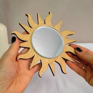 May include: A wooden sun-shaped mirror with a round, reflective surface. The mirror is light brown with a dark brown outline.