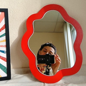 May include: A red, flower-shaped mirror with a wavy edge. The mirror is reflecting a person taking a selfie with a smartphone.