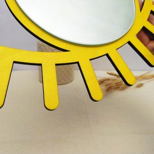 K&ouml;nnte beinhalten: Ein leuchtend gelber Wandspiegel mit einer kreisf&ouml;rmigen Spiegelfl&auml;che, umgeben von einem Rahmen aus strahlenf&ouml;rmig angeordneten, flachen, rechteckigen Holzlamellen, die an stilisierte Wimpern oder Sonnenstrahlen mit schwarzen Kanten erinnern.