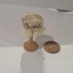 May include: A miniature doll bonnet, with a striped, sheer crown and a delicate lace trim. The bonnet is displayed on a small wooden stand. A penny is next to the bonnet for size reference.