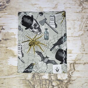 May include: A light gray Halloween-themed fabric with a repeating pattern of skulls, spiders, bats, axes, tombstones, and crows. The fabric has a white scalloped edge and a small white tag with a corn graphic.