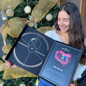 Può includere: Una scatola di presentazione nera con un design dorato e un certificato incorniciato. Il certificato presenta una nebulosa a forma di cuore e il nome "Marianne". Sullo sfondo si vede un albero di Natale decorato.
