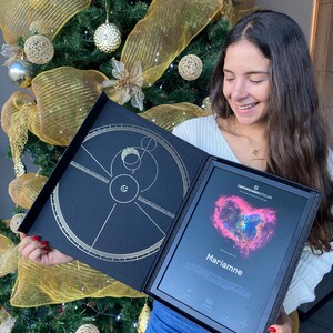 Può includere: Una scatola di presentazione nera con un design dorato e un certificato incorniciato. Il certificato presenta una nebulosa a forma di cuore e il nome "Marianne". Sullo sfondo si vede un albero di Natale decorato.