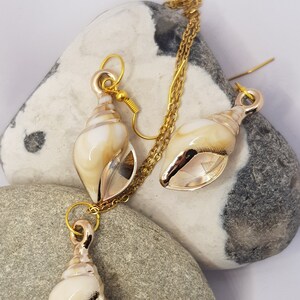 May include: A gold necklace and earrings set featuring a seashell design. The necklace has a delicate chain and a seashell pendant. The earrings are drop earrings with seashell charms.