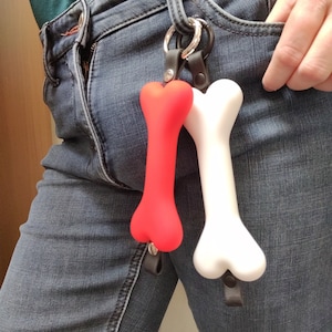 May include: A red and white dog bone-shaped keyring hanging from a silver ring. The keyrings are attached to a black leather strap. The person is wearing blue denim jeans.