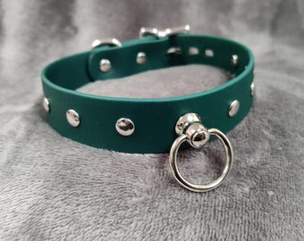 Könnte beinhalten: Ein dunkelgrüner Halsband mit silberfarbenen Nieten und einem großen Metallring. Das Halsband hat einen Schnallenverschluss und besteht aus einem glatten, einfarbigen Material. Es ist für das Tragen um den Hals konzipiert.
