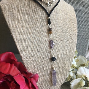 May include: A long, layered necklace with a black suede cord and silver accents. The necklace features a variety of gemstones, including amethyst, tiger's eye, and lava rock.