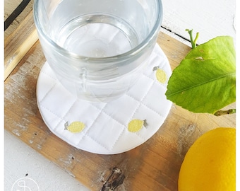 May include: White quilted coaster with embroidered yellow lemons, holding a clear glass of water. The coaster sits on a wooden tray, with a lemon and leaf in the background. A refreshing and decorative item.