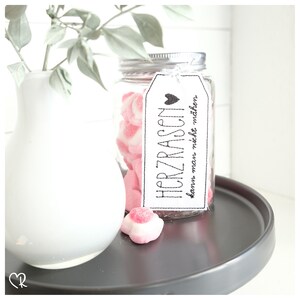 Peut inclure: Un bocal en verre rempli de bonbons en forme de cœur roses et blancs. Une étiquette blanche avec du texte noir indique "HERZRASEN kann man nicht machen".