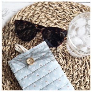 Puede incluir: Gafas de sol carey sobre un estuche acolchado azul claro con cierre de botón. El estuche tiene un estampado floral y está junto a un vaso de agua con cubitos de hielo. Los objetos están sobre una esterilla tejida.