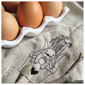 May include: A white egg carton with brown eggs and a linen fabric with an embroidered design of a bunny in a pot with a heart.