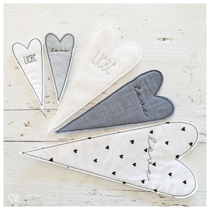 Puede incluir: Cinco decoraciones de pared en forma de corazón de tela con diferentes patrones y las palabras "love" y "liebe" bordadas en ellas. Los corazones están dispuestos en forma de abanico sobre un fondo de madera blanco.