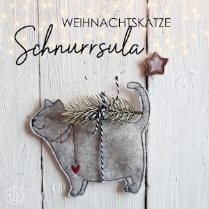 Könnte beinhalten: Ein grauer Filzkatzen-Anhänger mit einem kleinen roten Herzen auf der Brust. Die Katze trägt einen kleinen Tannenzweig auf dem Rücken und ist mit einer goldenen Perlenkette verziert. Der Anhänger ist mit "Weihnachtskatze Schnurrsulal" beschriftet und hat einen goldenen Stern oben drauf.