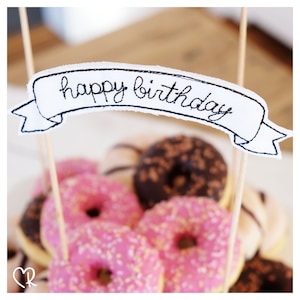 Peut inclure: Une bannière blanche avec des broderies noires qui disent "joyeux anniversaire" est placée au sommet d'une tour de beignets. Les beignets sont glacés et saupoudrés de pépites colorées.