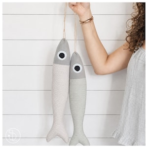 Könnte beinhalten: Zwei hängende fischförmige Stoffornamente, die an dünner Schnur befestigt sind. Ein Fisch hat einen hellbeigen Körper mit weißen Punkten und einen einfarbig grauen Kopf. Der andere Fisch hat einen grau-weiß gestreiften Körper und einen grauen Kopf.