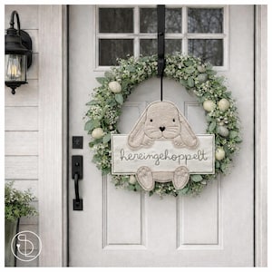 Puede incluir: Una corona de primavera con un conejito y el texto "hereingehoppelt" colgada en una puerta blanca. La corona está hecha de vegetación, pequeñas flores blancas y huevos moteados. El conejito es de color marrón claro y sostiene un letrero.