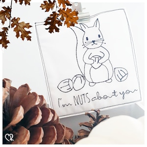 May include: A white fabric with black embroidery of a squirrel holding a nut and the text "I'm NUTS about you".