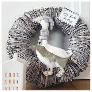 May include: A woven twig wreath with a plush goose decoration. The goose wears a grey scarf and hat, with white feathers and green leaf details. A sign reads "Gans frohe Feiertage".
