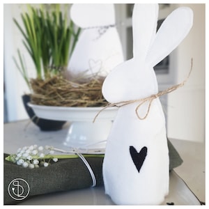 Puede incluir: Conejo de fieltro blanco con un corazón negro en el pecho. El conejo está atado con una cuerda y está sentado en una mesa con una servilleta verde y un ramo de flores blancas.