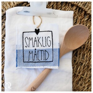 Könnte beinhalten: Ein weißes Küchentuch mit einem blau-weiß karierten Rand. Ein kleines besticktes Schild mit einem schwarzen Herzen und dem Text "SMAKLIG MÅLTID" ist an dem Tuch befestigt. Ein Holzlöffel liegt auf dem Tuch.