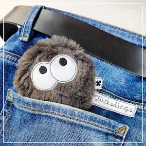 Könnte beinhalten: Ein graues, flauschiges Spielzeug mit großen weißen Augen und schwarzen Pupillen steckt in der Tasche einer blauen Jeans. Die Jeans hat einen braunen Ledergürtel und ein weißes Etikett mit der Aufschrift "Glücksdings".