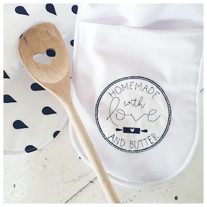 Op de afbeelding: Een witte ovenwant met een zwart cirkelvormig ontwerp met de tekst "Homemade with love and butter". Op de voorgrond ligt een houten lepel met een hartvormige uitsparing.