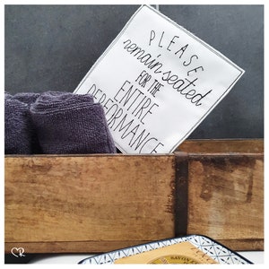 Könnte beinhalten: Ein weißes Schild mit schwarzem Text, auf dem "Bitte bleiben Sie für die gesamte Vorstellung sitzen" steht, lehnt an einer Holzkiste. Die Kiste ist mit grauen Handtüchern gefüllt.