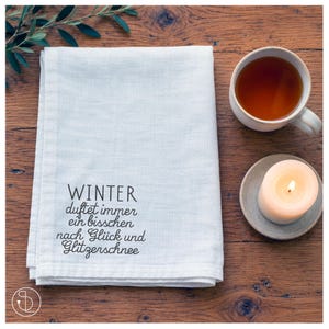 May include: A white rectangular linen napkin featuring black printed German text that reads: WINTER duftet immer ein bisschen nach Glück und Glitzerschnee. The fabric has a visible woven texture and is folded neatly on a dark wooden surface.