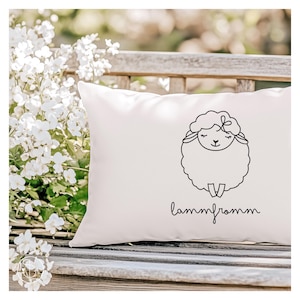May include: A white rectangular cushion featuring a black line drawing of a sheep and the word "lammfromm" in script. The sheep has a fluffy body and a smiling face. The cushion is on a wooden bench with white flowers in the background.