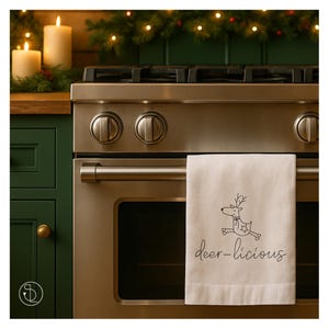 May include: A white kitchen towel with a black outline drawing of a reindeer and the text "deer-licious". The towel hangs on a stainless steel oven. The background includes a green cabinet, candles, and Christmas greenery.
