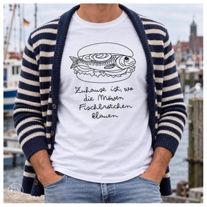 Könnte beinhalten: Ein weißes T-Shirt mit einer schwarzen Strichzeichnung eines Fischbrötchens und dem Text 'Zuhause ist, wo die Möwen Fischbrötchen klauen'. Das T-Shirt wird unter einer dunkelblau-weiß gestreiften Strickjacke mit Knöpfen getragen.