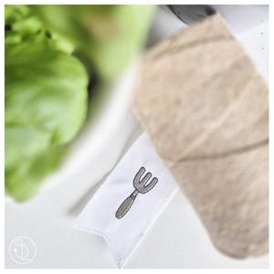 May include: A white fabric tag with a gray and brown embroidered garden fork design. A roll of beige twine is visible in the background, along with a blurred green plant. The tag is likely for a gardening-themed product.