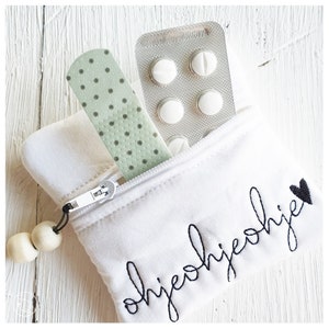 May include: White zippered pouch with the word "ohje ohje" embroidered in black with a small black heart. The pouch contains a green polka dot bandage and a blister pack of white pills.