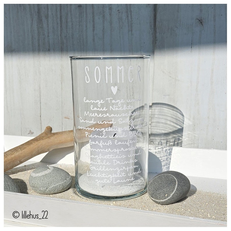 May include: A clear glass candle holder with the word "SOMMER" and a heart etched on the front. The candle holder is filled with white sand and has a white inscription that reads "lange Tage und laue N&auml;chte, Meeresrauschen, Sand und Sonne, summengek&uuml;sster, Picnic und BBQ, barfu&szlig; laufen, Sommersprossen, Spaghetti Eis und k&uuml;hle Drinks, Grillenzirpen, Leichtigkeit und gute Laune."