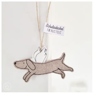 Könnte beinhalten: Ein Filz-Dackel-Ornament in Hellbraun mit schwarzen Nahtdetails und weißen Flügeln. Ein kleines Etikett mit dem Text "Schutzdackel" ist angebracht. Das Ornament hängt an einer Schnur.