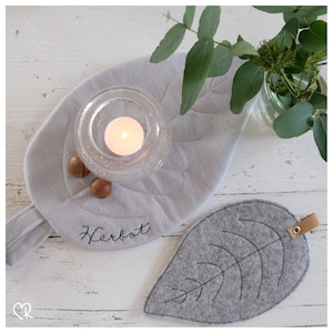 Peut inclure: Un dessous de plat en forme de feuille en feutre gris avec le mot "Herbst" brodé dessus. Un bougeoir en verre avec une bougie allumée est posé sur le dessous de plat. Il y a deux petites noix brunes sur le dessous de plat.