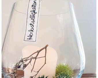 Può includere: Un vaso di vetro trasparente con un'etichetta bianca che dice "Herbstlich Garten" e una candela bianca all'interno. Il vaso è pieno di rami secchi e piante verdi.