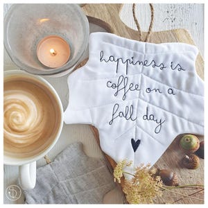 May include: White fabric leaf-shaped ornament with the text "happiness is coffee on a fall day" embroidered in black thread. A small black heart is embroidered below the text. The ornament is hanging from a brown twine string.