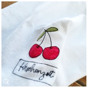 Könnte beinhalten: Weißer Stoff mit gestickten roten Kirschen und grünen Stielen. Der Stoff hat den Text "Kirschenzeit" in Schwarz gestickt.