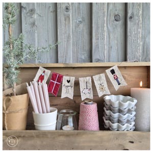 Könnte beinhalten: Ein rustikales Holzregal zeigt festliche Weihnachtsdekorationen. Ein Banner mit gestickten Designs hängt über Kerzen, einer Topfpflanze und einem Stapel weißer Keramikschalen. Das Banner zeigt einen Vogel, eine Flagge und andere weihnachtliche Motive.